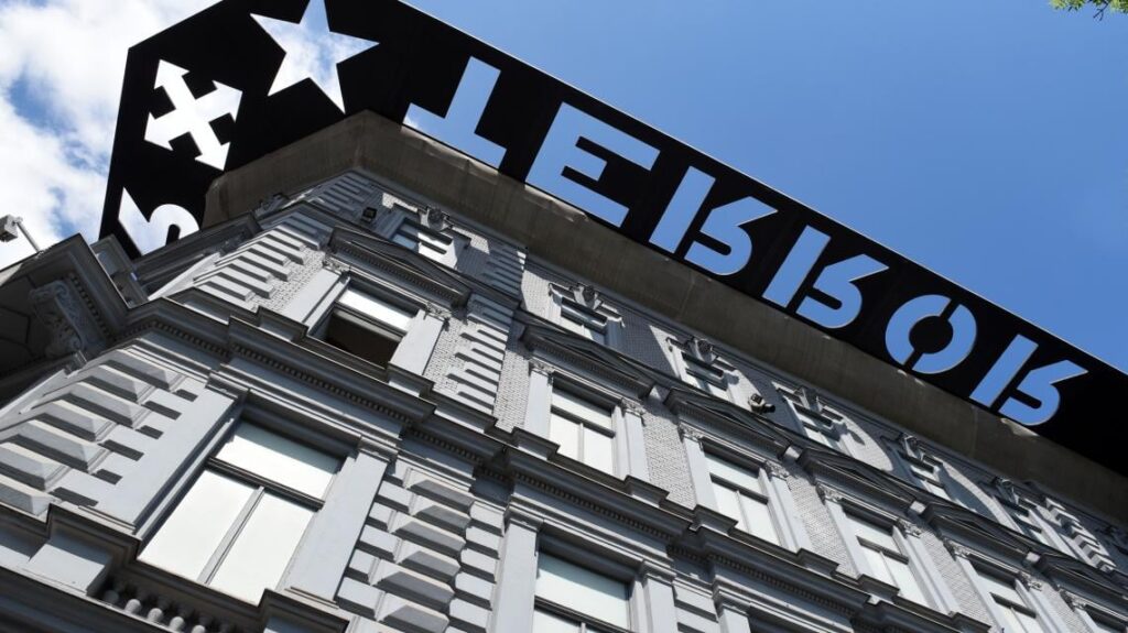Ornate multi-story building with cut-out symbols on the roof, viewed from a low angle against blue sky.