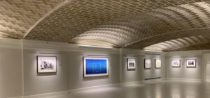 Art gallery with vaulted patterned ceiling and framed artworks evenly spaced on neutral walls.