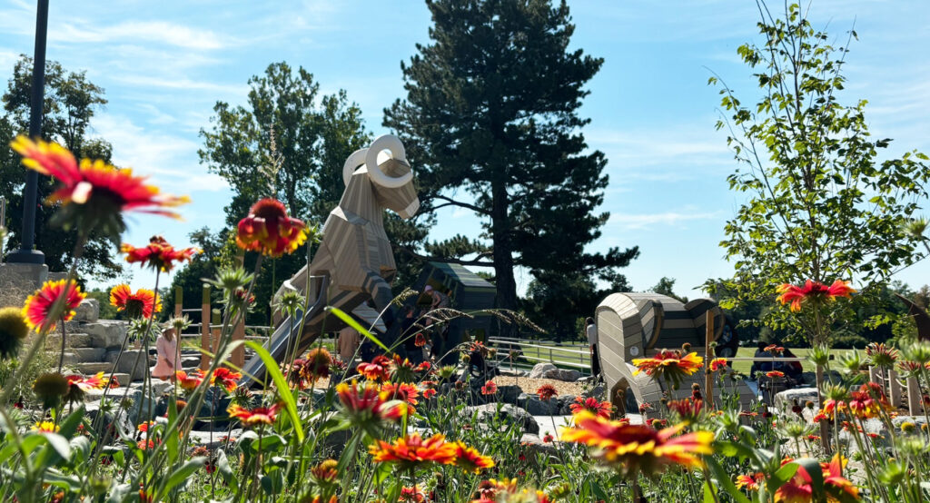 Garden with red, yellow flowers, wooden animal sculptures, trees, and blue sky backdrop.