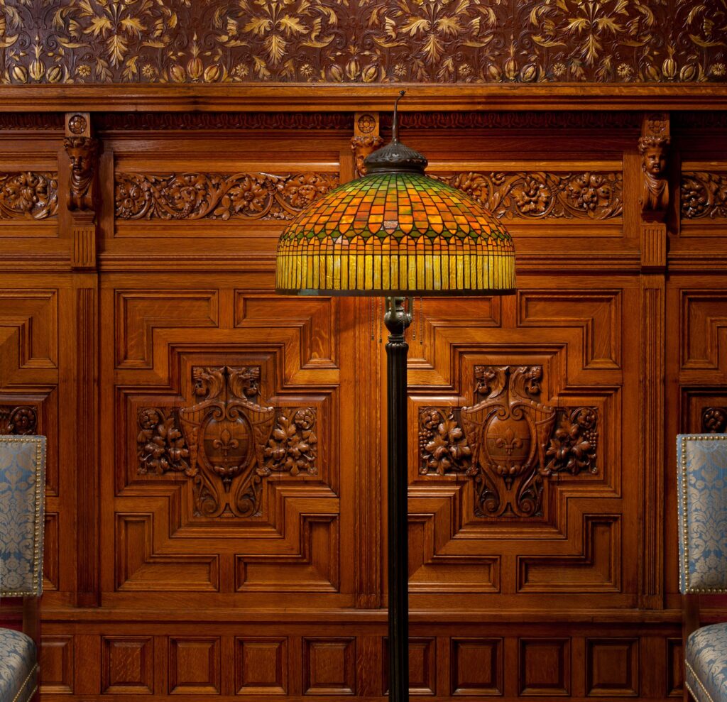 Ornate carved wooden wall, stained glass lamp, and blue upholstered chairs with nailhead trim.