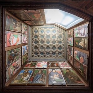Decorated room with geometric ceiling and framed paintings of figures holding books or scrolls.