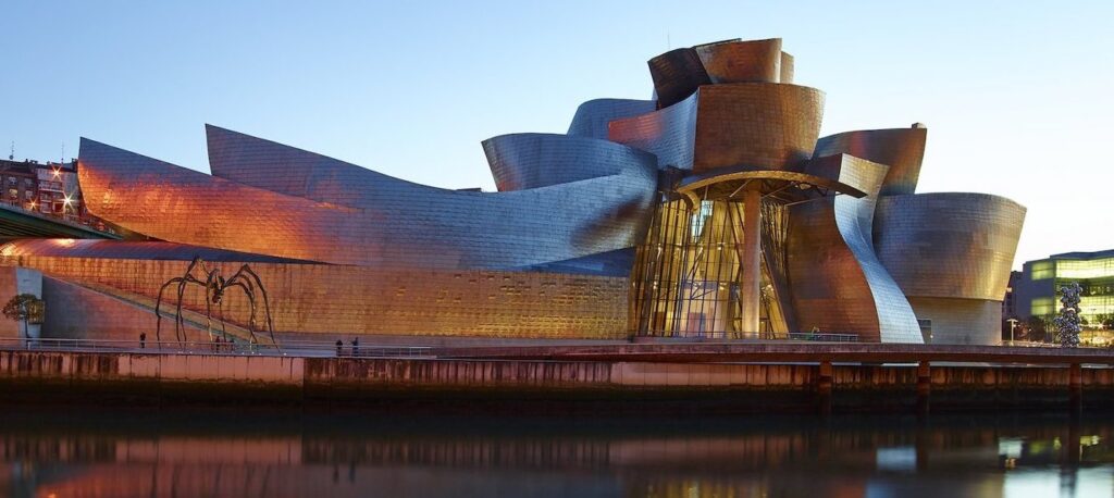 Modern building with reflective surfaces by water, large spider sculpture nearby.