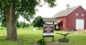 Large red barn with amenities sign, trees, and historical plow on grassy area.
