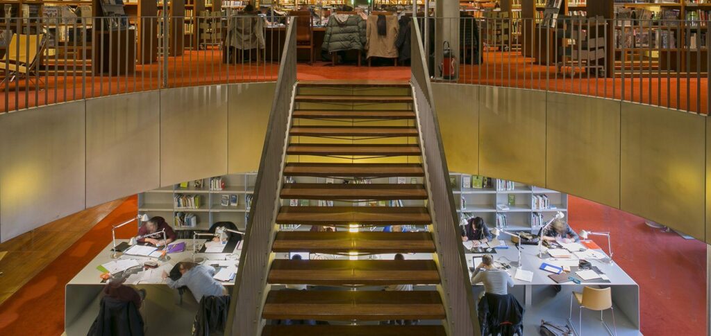 Library interior with central staircase, study areas, and bookshelves on two levels.
