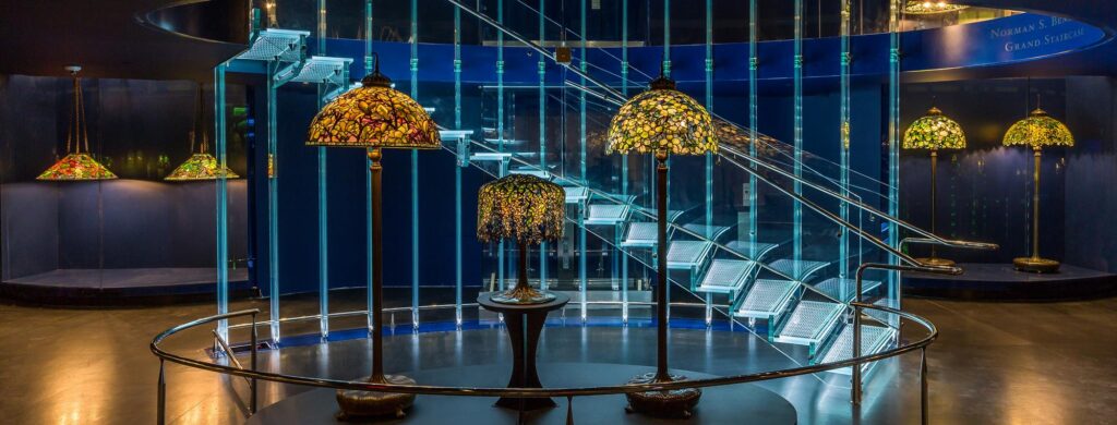 Stained glass lamps on stands in a dim space with a modern metal and glass staircase background.