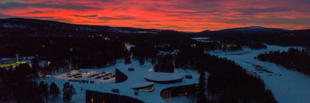 Aerial view of a snowy landscape, modern building, and vivid sunset with forested hills.