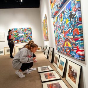 People examining artworks in a brightly lit gallery, one taking notes and the other viewing photos.