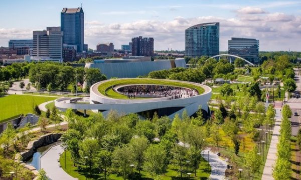 Modern urban landscape with a circular building, lush green park, high-rise buildings, and bridge.